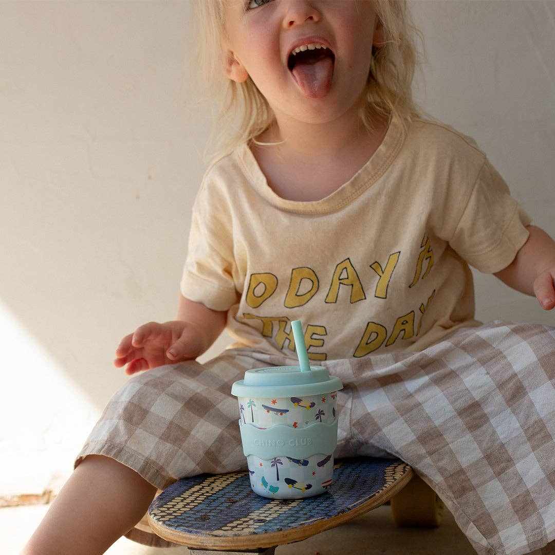 Child sitting on skateboard holding an 8oz Skateboard Kids Keep Cup with straw and colorful skateboard design