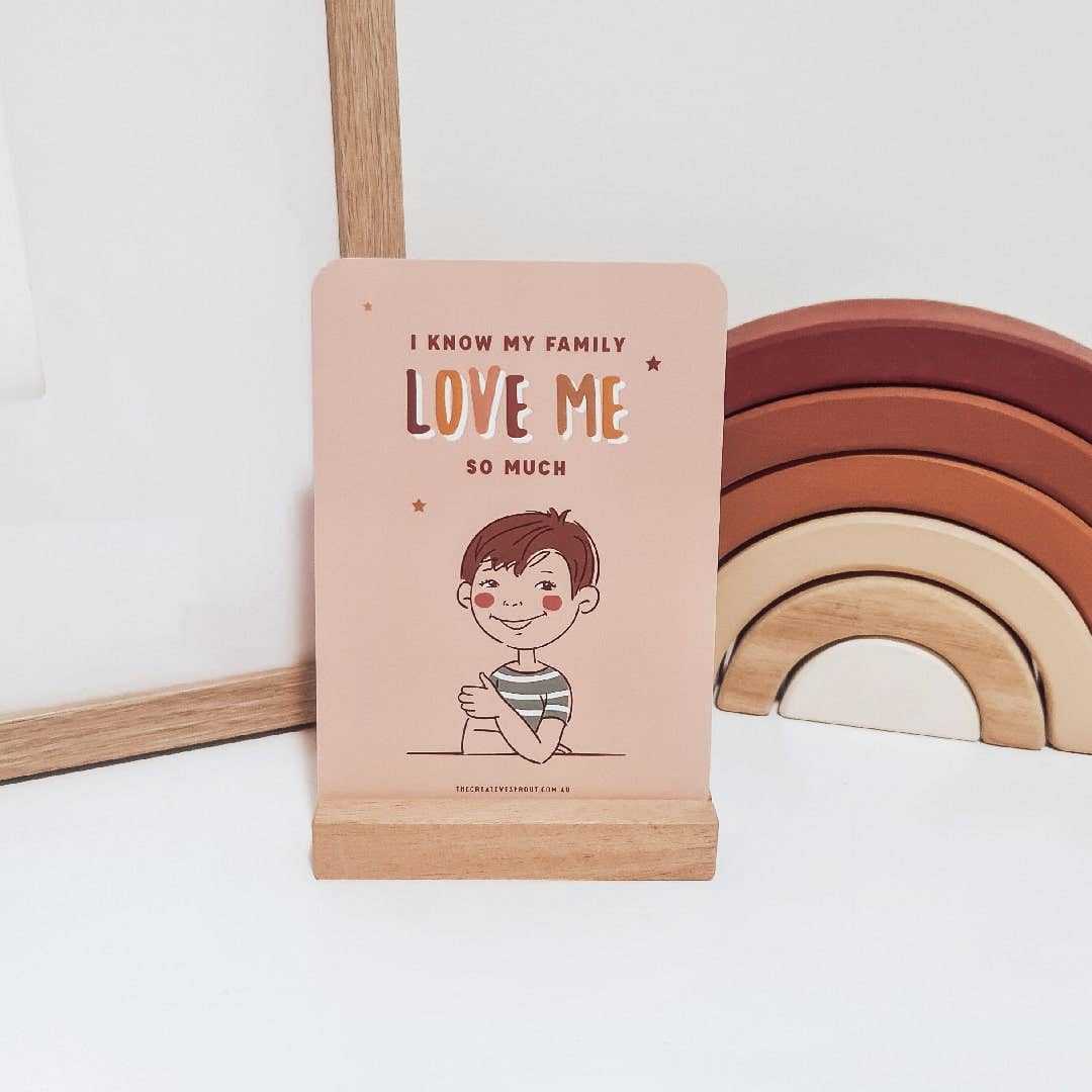 Kids Confidence Cards featuring an illustrated affirmation card with the message "I know my family love me so much" on a wooden stand with a rainbow toy in the background