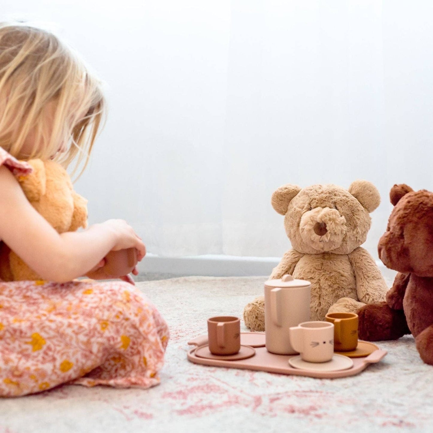 Child playing with Maple the Bear soft toy and tea set on carpet in bright room
