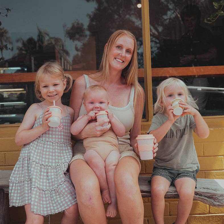 Mother sitting with three children holding pink daisy babychino cups and drinking beverages outside cafe