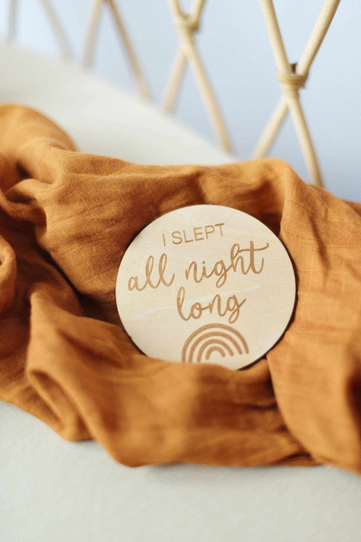 Kiin Baby Wooden Milestone Disc etched with "I slept all night long" text and rainbow design on a soft brown cloth background