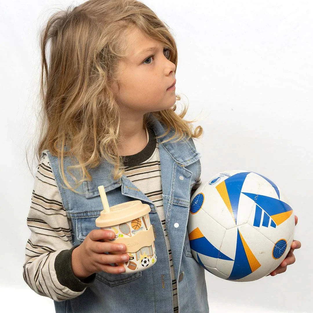 Child holding a sports-themed 4oz bamboo babychino cup with silicone lid and grip band next to a blue and orange soccer ball