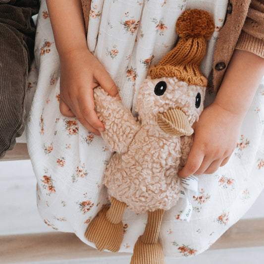 A plush toy duck with a knitted beanie being held by a child sitting on a lap with a floral dress.