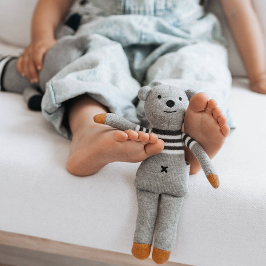 A small gray and white striped teddy bear with a marble pattern, holding a child's foot, while the child sits on a bed.