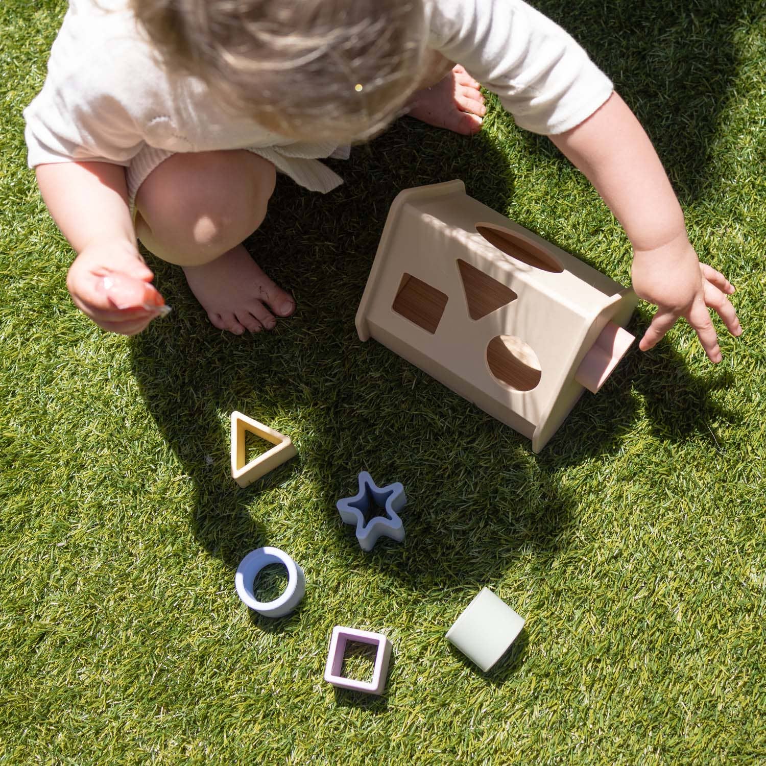 A baby playing with a silicone shape sorting set on grass, with sorters in various shapes and colors scattered around.