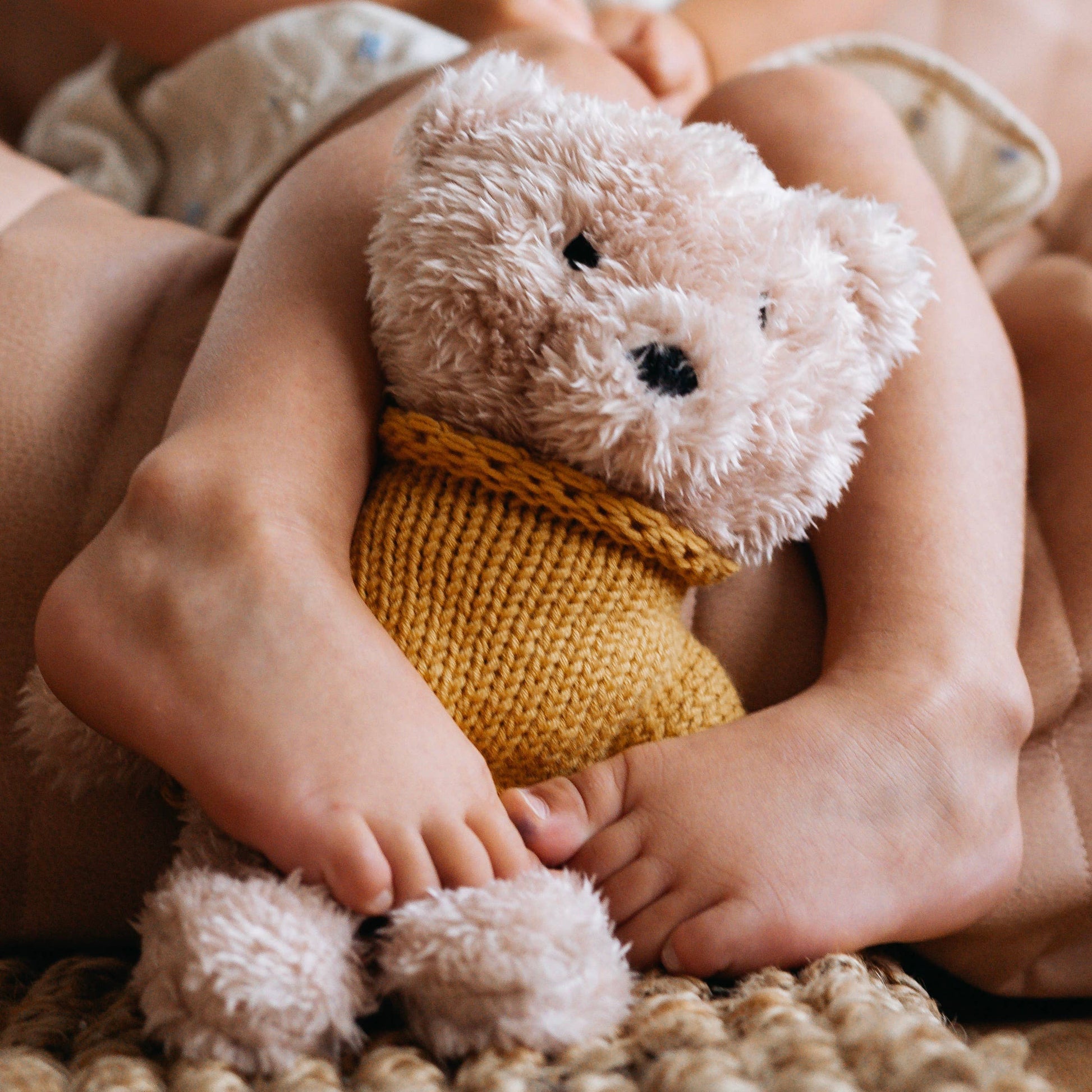 A child holding a beige plush toy bear with a yellow knitted outfit.